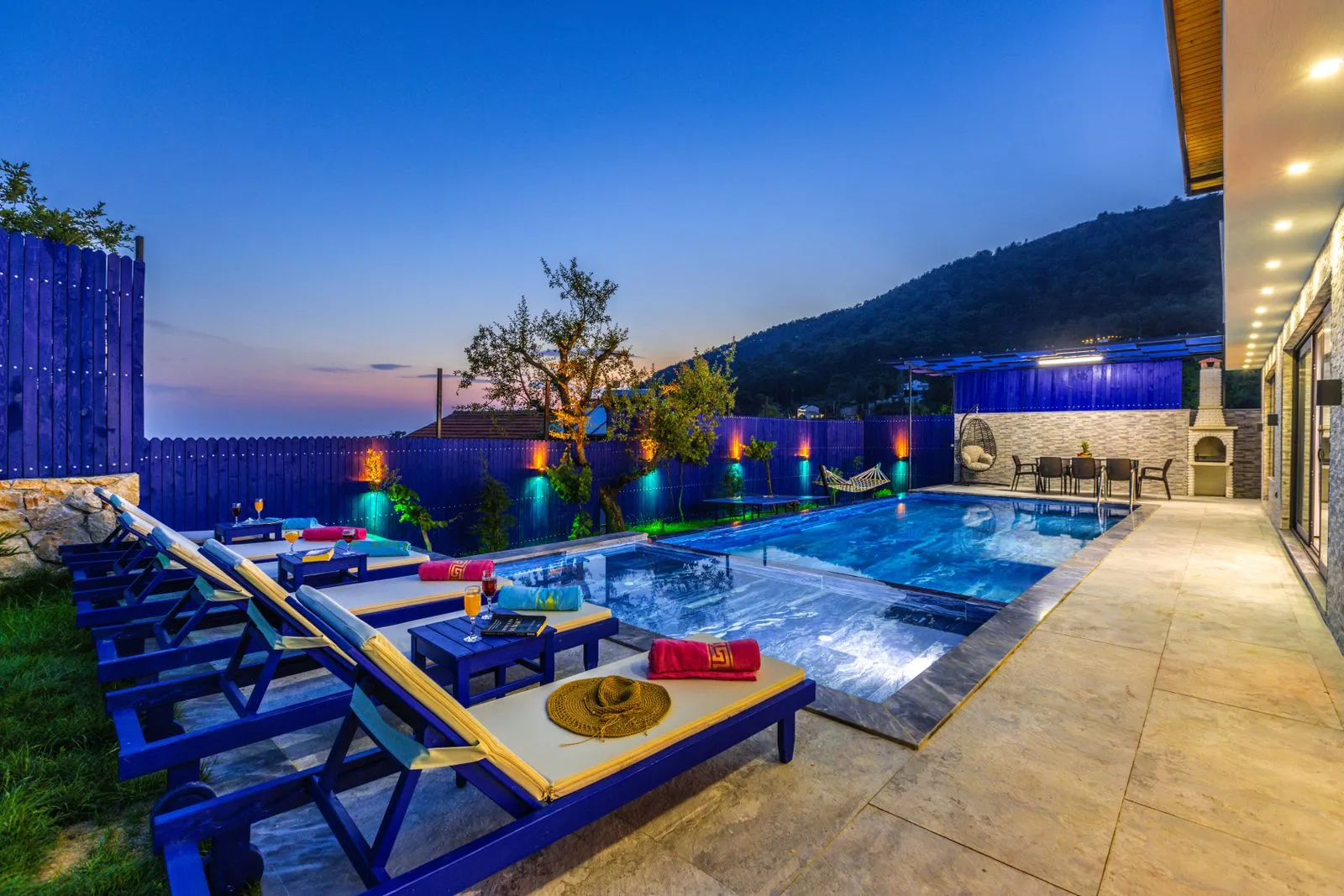 A backyard pool at dusk with travertine decking, lounge chairs, and warm lights