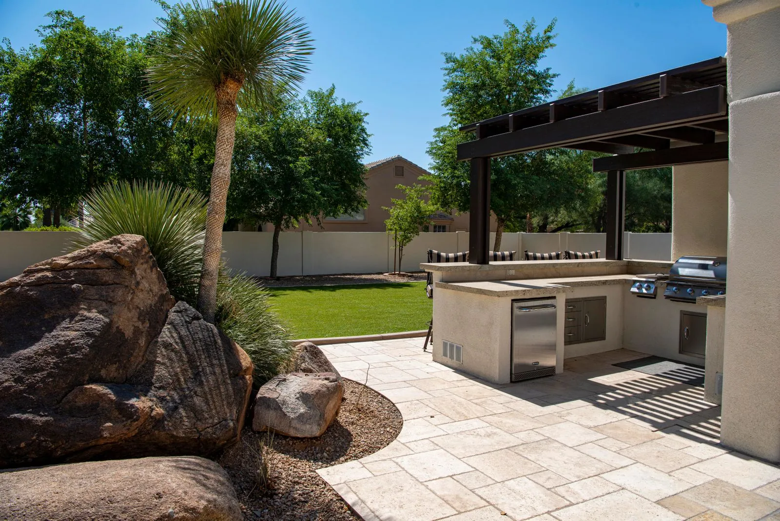 An outdoor kitchen under a pergola with travertine pavers and desert boulders